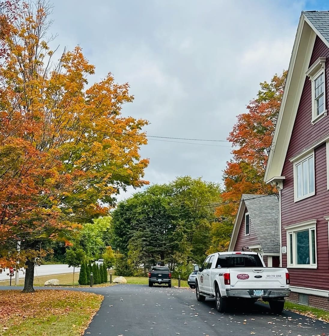 Driveway scene in Seacoast NH with fall foliage, red home exterior, parked trucks, and overcast sky