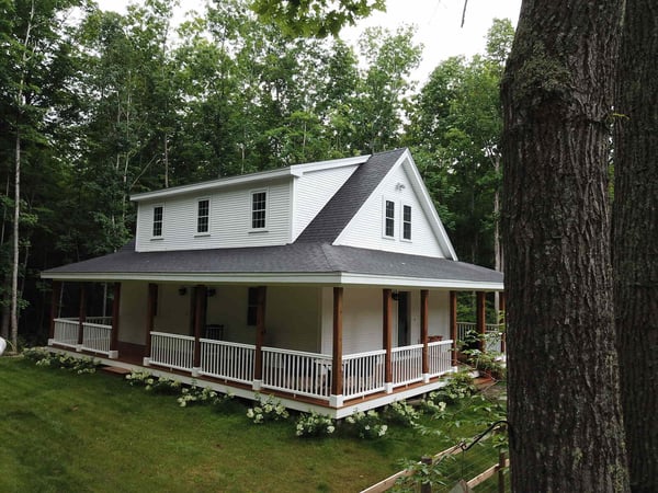 Aerial view of a custom home with a wraparound porch, surrounded by forested landscape, built by Oxland Builders in New Hampshire