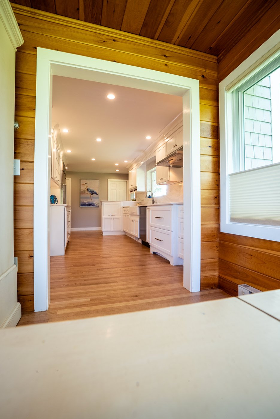 View into white kitchen with wood floors from sunroom, a remodel by Oxland Builders in New Hampshire