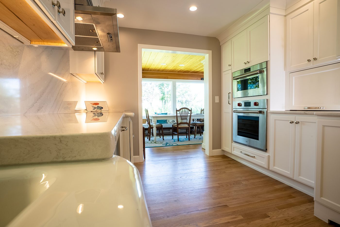 White kitchen in New Hampshire with marble counters, stainless steel oven, and a view into a sunroom with wood ceiling