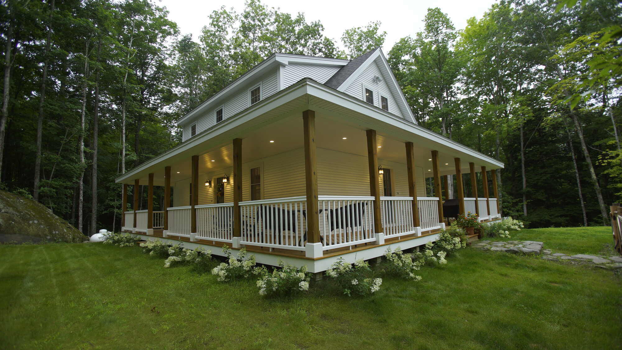 Custom home with a wraparound porch surrounded by lush greenery, built by Oxland Builders in New Hampshire