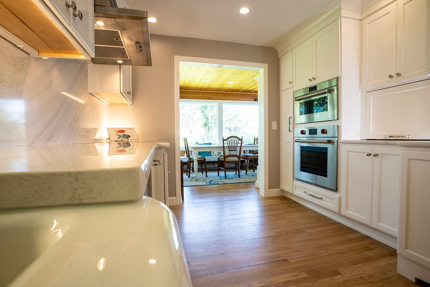 White kitchen in New Hampshire with marble counters, stainless steel oven, and a view into a sunroom with wood ceiling White kitchen in New Hampshire with marble counters, stainless steel oven, and a view into a sunroom with wood ceiling